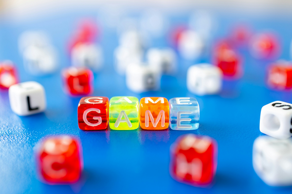Bunte Buchstabenwürfel bilden das Wort “GAME” auf einem blauen Tisch, weitere Würfel mit Buchstaben liegen unscharf im Hintergrund.
