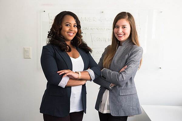 Zwei Frauen stehen lächelnd vor einem beschriebenen Whiteboard in einer Büroumgebung.