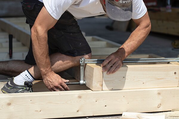 Ein Mann kniet auf Holzbalken, die mit einem Holzzwinger zusammengehalten werden.