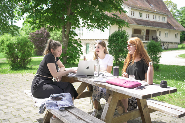 Drei Studentinnen sitzen an einer Holzbank im Grünen und arbeiten gemeinsam an einem Projekt.